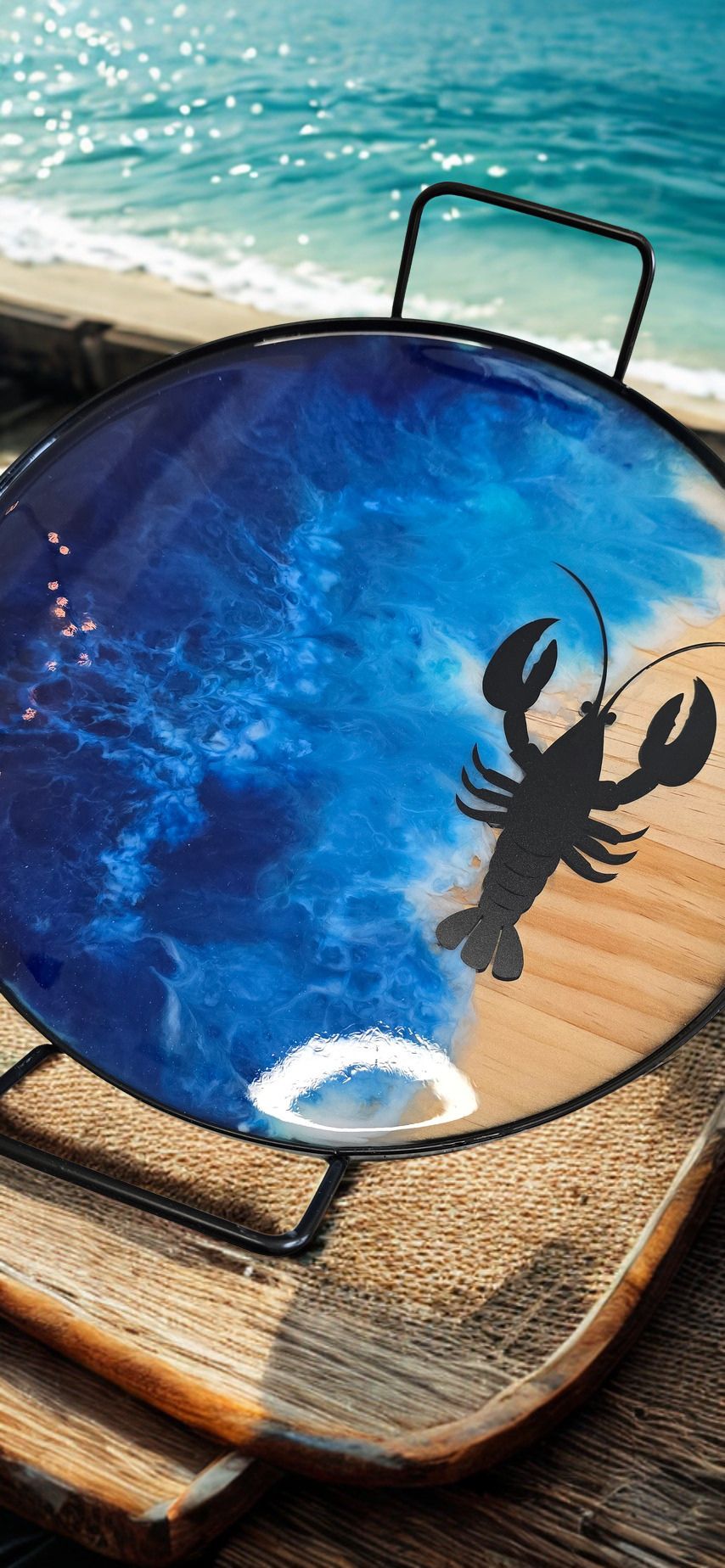 Round blue plate with a black silhouette of a lobster on a wooden surface by the ocean.